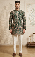 A man modeling a dark green embroidered kurta with intricate floral patterns, paired with off-white trousers and brown loafers, standing in a decorated indoor setting.